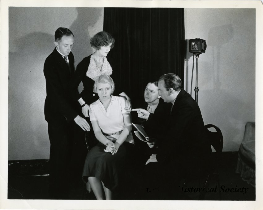 Black and white photo taken of a group of the Mummers, a group of radio actors and actresses, known for their horror series the Hermit's Cave, dramatically posed in the WJR studio. They are identified on a piece of note paper pasted to the verso with a "Public Address Systems, 340 East Grand Boulevard" header from left to right as Irving Butterfield, Prudence Butterfield, Donelda Currie, Charles Penman, and Bruce Myers. The Butterfields stand behind Currie, who sits gazing off toward the camera, as Myers, also seated, points at her accusingly, and Penman looks on holding the script. A WJR light stands next to a curtain hung in the background.